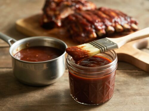 A jar of thick homemade barbecue sauce with a basting brush and glazed ribs behind it